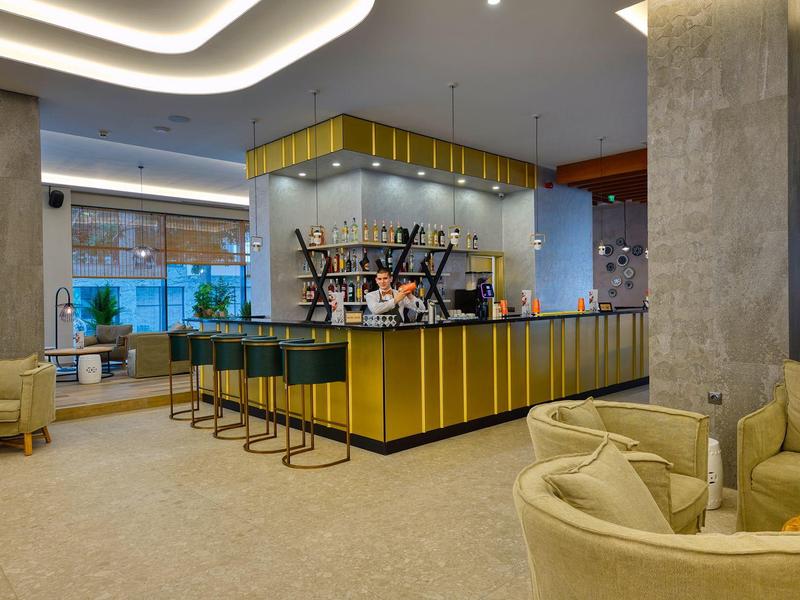 Moderní hotelový bar s žlutým barem a zelenými barovými židličkami ve stylovém prostředí.