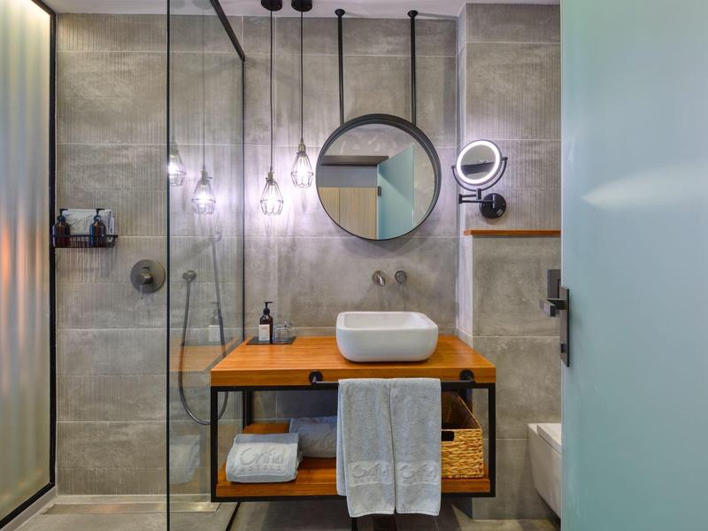 Moderní koupelna s tmavými dlaždicemi, dřevěnou umyvadlovou skříňkou a kulatým zrcadlem.