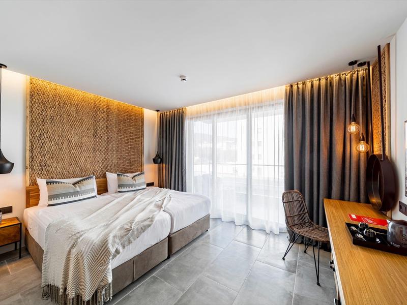 Moderní hotelový pokoj s manželskou postelí, velkými okny a elegantním interiérem.