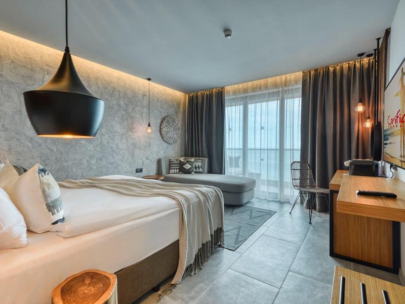 Moderní hotelový pokoj s manželskou postelí, posezením a velkým oknem se závěsy