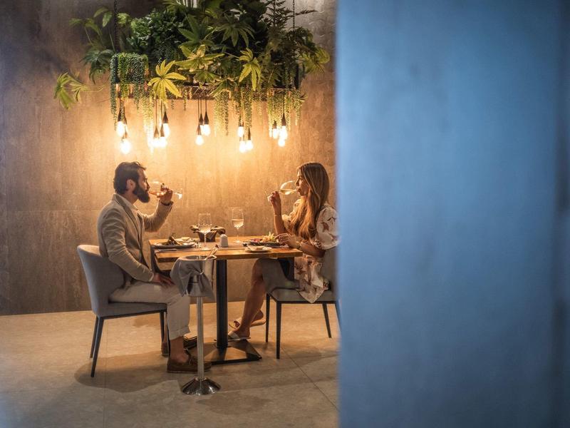 Pár sedí u stolu při svíčkách v útulné restauraci s závěsnými lampami a rostlinami.