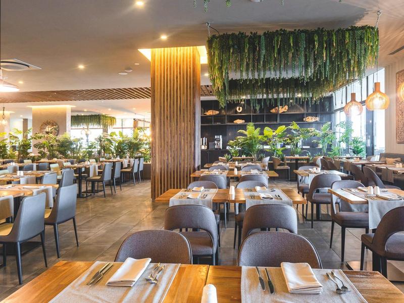 Moderní restaurace s dřevěnými stoly, židlemi a zelenými rostlinami v světlém prostředí.