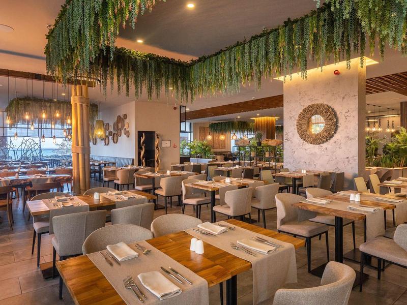 Moderně zařízená restaurace se dřevěnými stoly, pohodlnými židlemi a zelenými rostlinami.