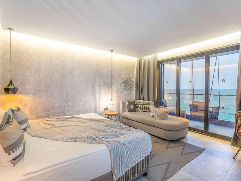Moderní hotelový pokoj s manželskou postelí, pohovkou a výhledem na moře přes velká skleněná okna.