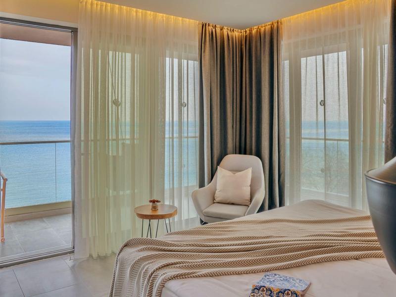 Světý hotelový pokoj s výhledem na moře, závěsy a pohodlná křesla u okna.