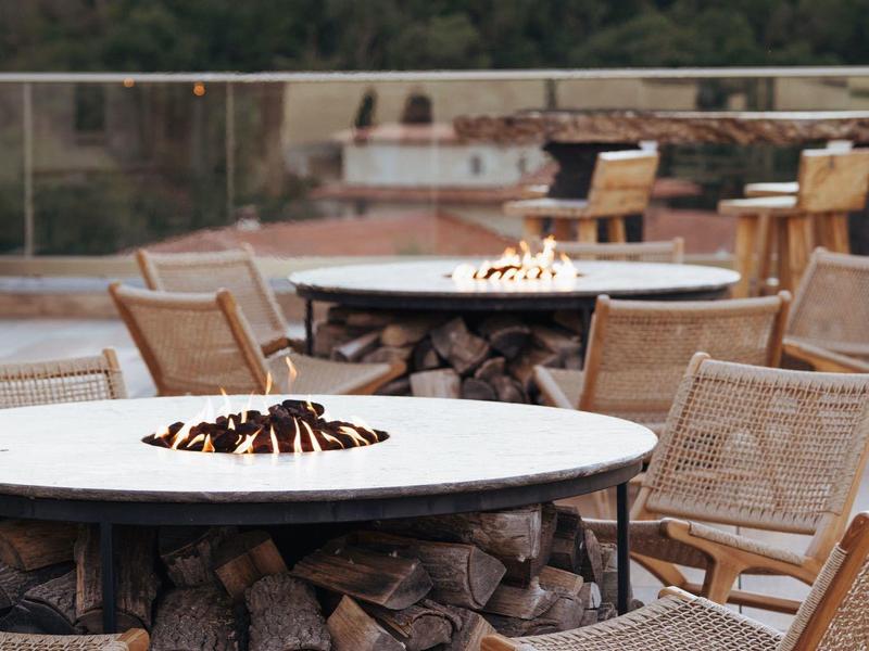 Moderní venkovní prostor s kulatými ohništi a dřevěným nábytkem před lesnatou krajinou.