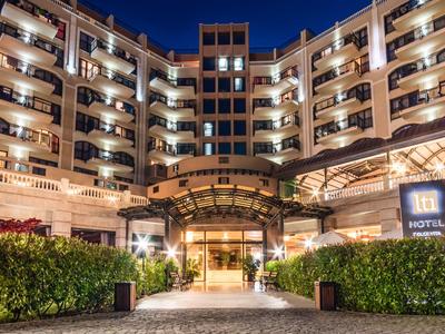 Großes Hotelgebäude mit vielen beleuchteten Balkonen und Eingang, umgeben von grünen Pflanzen.