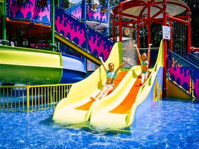 Bunte Wasserrutschen mit zwei gelben Röhren ins blaue Planschbecken, umgeben von buntem Gelände.