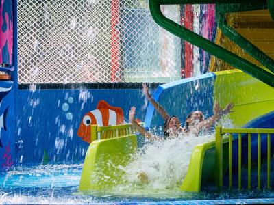 Kinder rutschen eine grüne Wasserrutsche in einem bunten Schwimmbad mit Cartoon-Fischen.