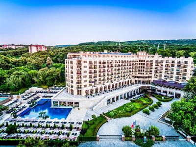 Großes Hotel mit Pool, Liegestühlen und viel Grün in einer ruhigen Landschaft unter blauem Himmel.