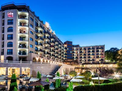 Modernes Hotelgebäude mit Balkonen, beleuchtet bei blauem Abendhimmel und gepflegtem Garten.