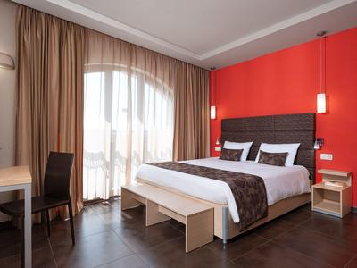 Modern hotel room with large bed, red accent wall, and floor-to-ceiling windows with curtains.