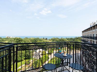 Balcony with table and chairs overlooking garden, sea, and hotel building under clear sky.