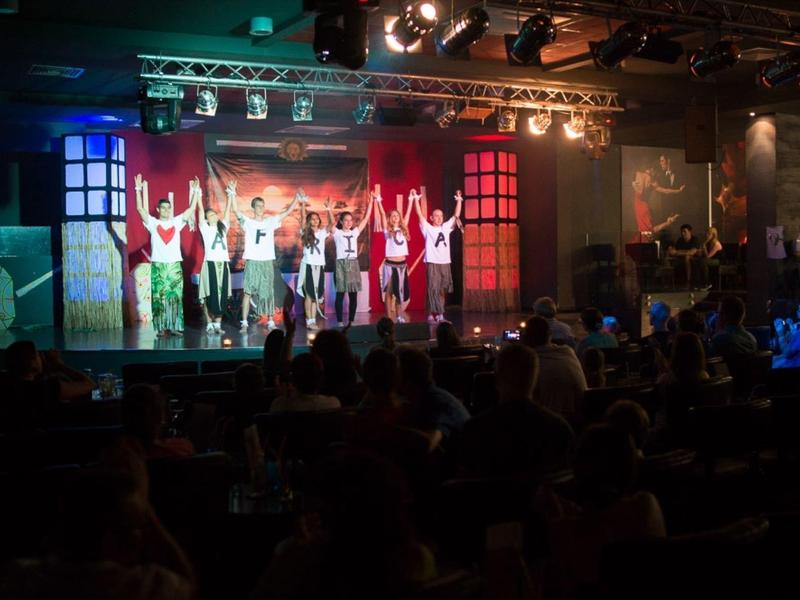 Eine Bühnenaufführung mit vielen Tänzern in roter und weißer Kleidung vor einem Publikum in einem Theater.