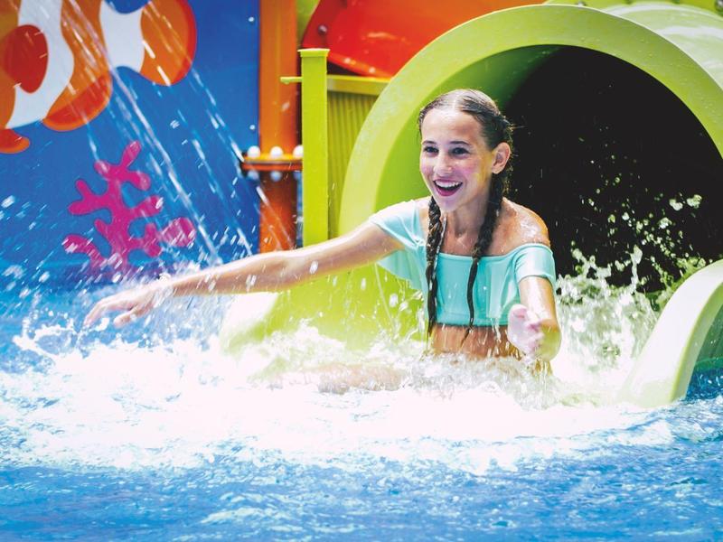 Glückliches Mädchen kommt aus einer grünen Wasserrutsche ins Schwimmbecken