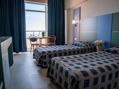 Zweibettzimmer mit blauen Vorhängen, zwei Sesseln am Fenster und geometrisch gemusterten Bettdecken.