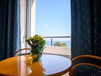 Blauer Himmel und Meerblick durch ein Fenster mit blauen Vorhängen, runder Tisch mit gelber Blume.