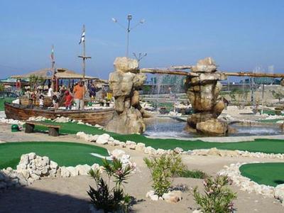 Minigolfplatz mit grünen Bahnen, Felsenhindernissen und Schiffen im Hintergrund am Hafen.