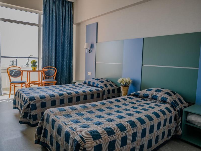 Zwei Einzelbetten mit blau karierten Decken, neben Fenster mit blauen Vorhängen und orangenen Stühlen.