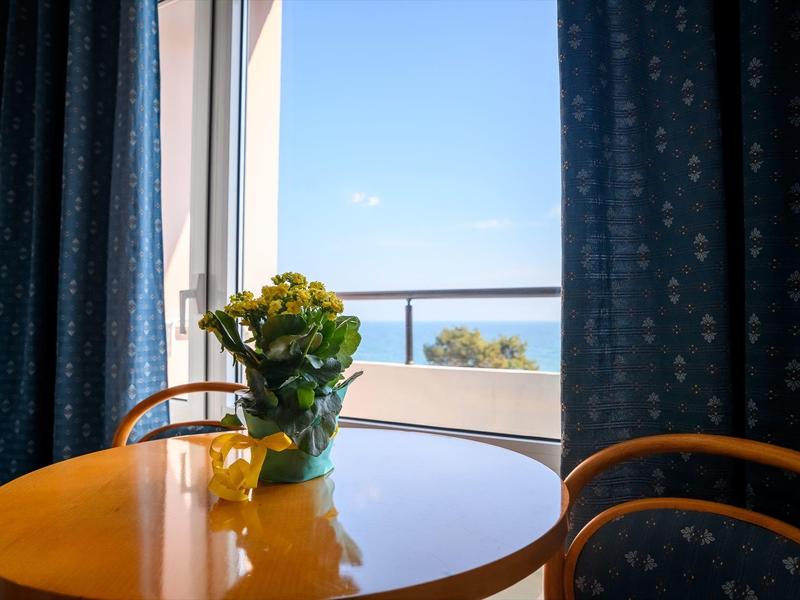 Blauer Himmel und Meerblick durch ein Fenster mit blauen Vorhängen, runder Tisch mit gelber Blume.