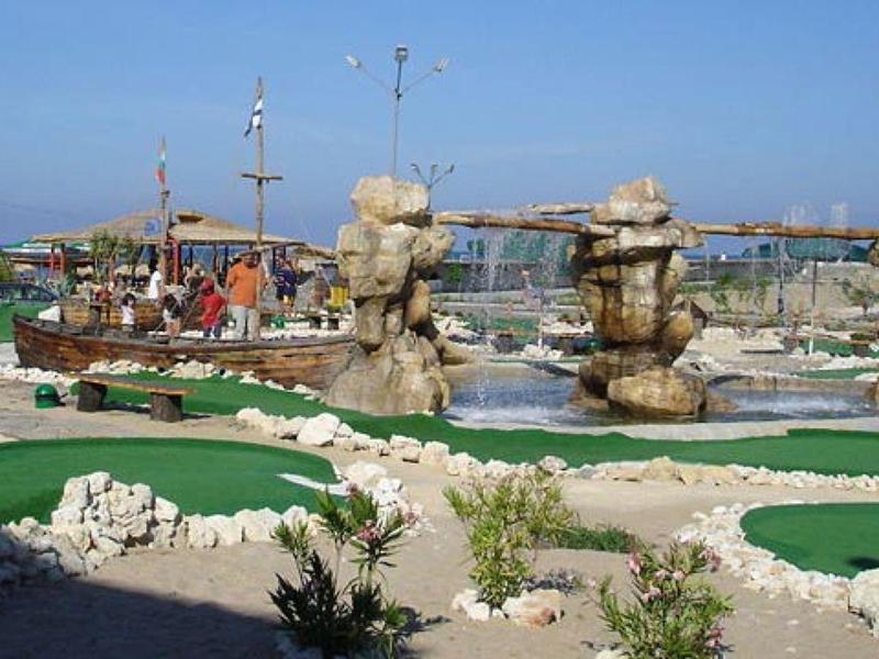 Minigolfplatz mit grünen Bahnen, Felsenhindernissen und Schiffen im Hintergrund am Hafen.