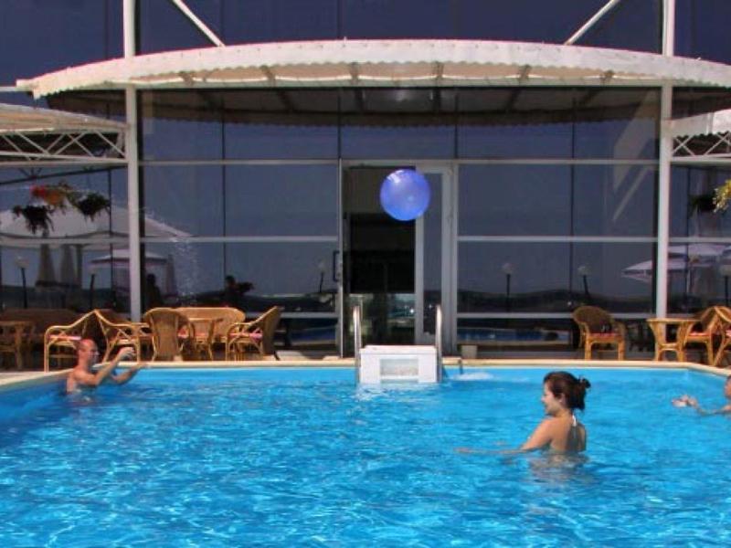 Schwimmbecken mit blauem Wasser, mehreren Menschen und weißen Liegestühlen vor Glasfassade.