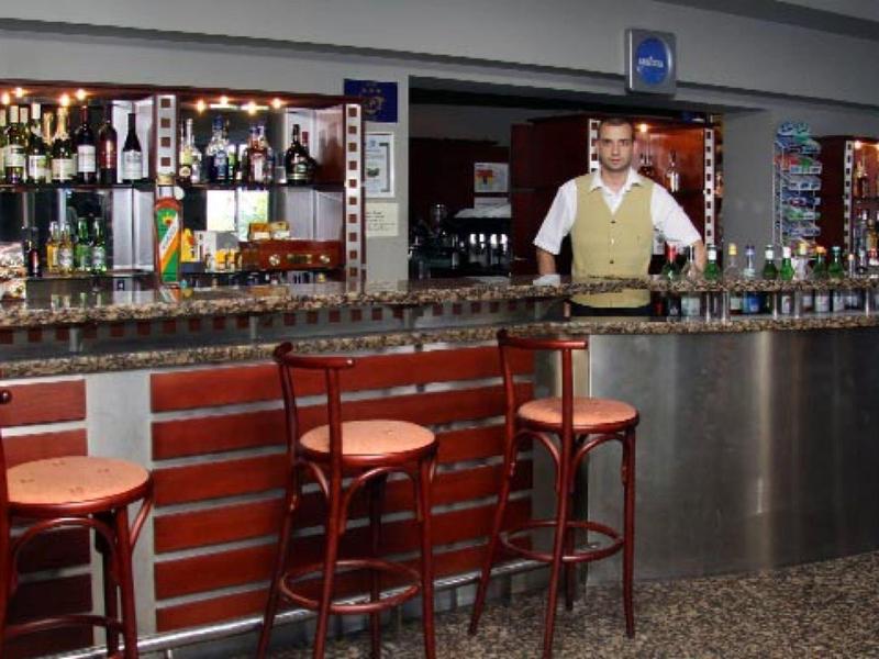 Bar mit drei Holzstühlen, Helm-Dekor und Barkeeper hinter langem Metall- und Granittresen.