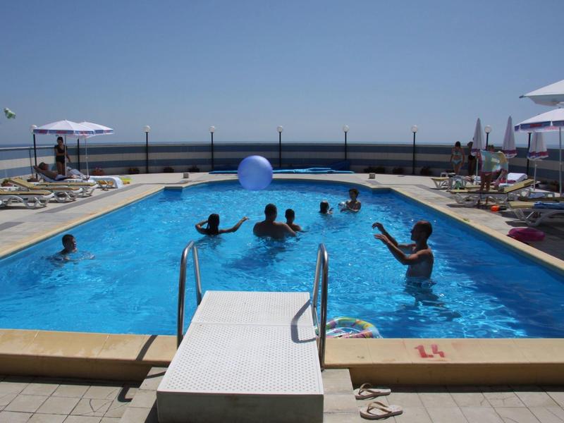 Freiluftpool mit mehreren Personen, blauer Himmel, Liegestühle und Sonnenschirme am Beckenrand.