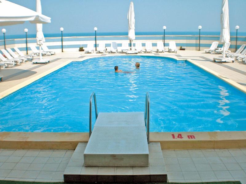 Blauer Swimmingpool mit zwei Personen, Liegestühlen und Meerblick im Hintergrund.