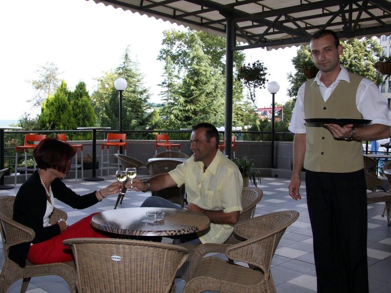 Zwei Gäste sitzen an einem Tisch auf einer Terrasse, ein Kellner steht mit Menü daneben.