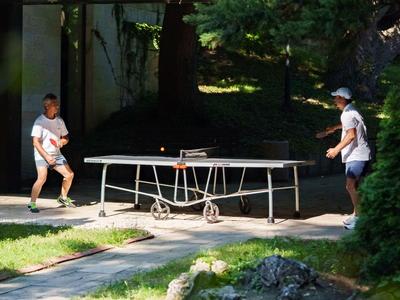 Deux personnes jouent au tennis de table dans un jardin d'hôtel sous des arbres.