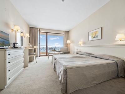 Bright hotel room with two beds, TV, desk, and balcony with sea view.