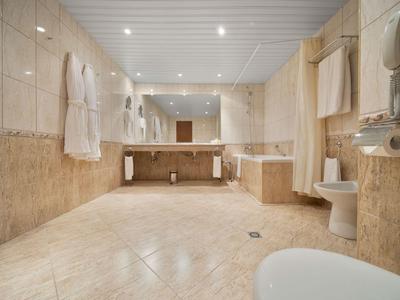 Grande salle de bain avec carrelage clair, deux lavabos, toilettes et peignoirs accrochés au mur.