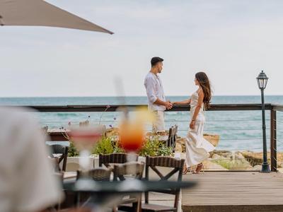 Un couple marche le long d'un chemin du port avec vue sur la mer et des tables au premier plan.