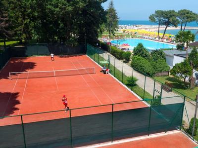 Vue d'un court de tennis rouge à côté d'une plage avec parasols et arbres.