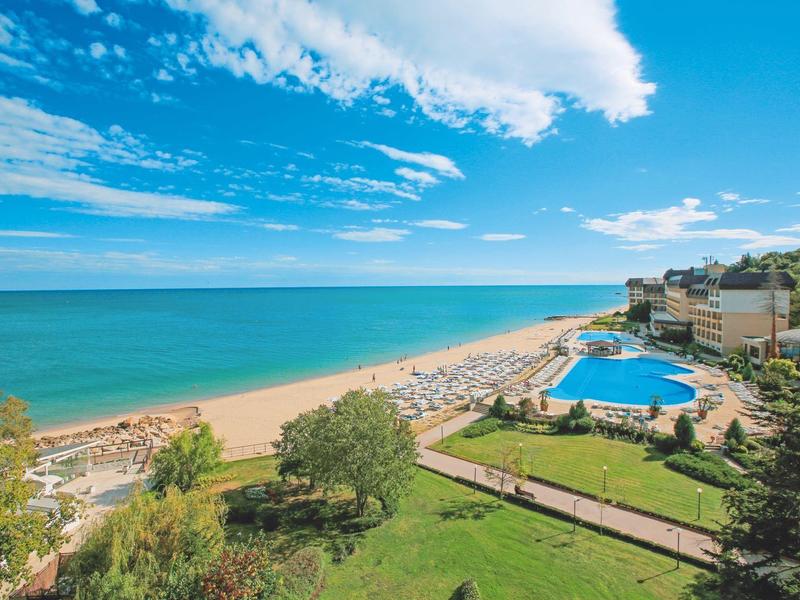 Beach with blue pool and sun umbrellas next to a hotel under a blue sky.