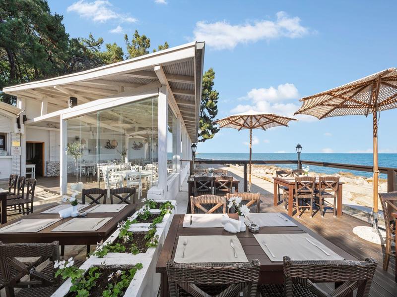 Modern beachside restaurant with wooden furniture, umbrellas, and sea view under clear skies.