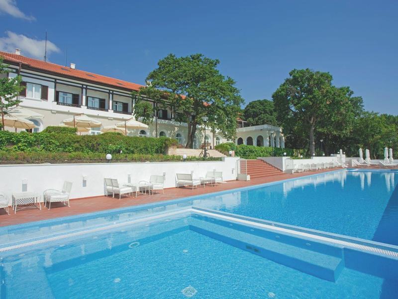 A large outdoor pool in front of a hotel with white walls and red roofs under a clear blue sky.