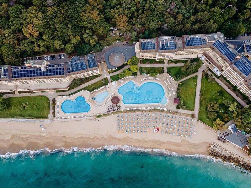 Aerial view of a hotel with pools on the beach and forest background.