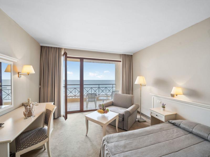 Bright hotel room with double bed, seating area, and balcony overlooking the sea.