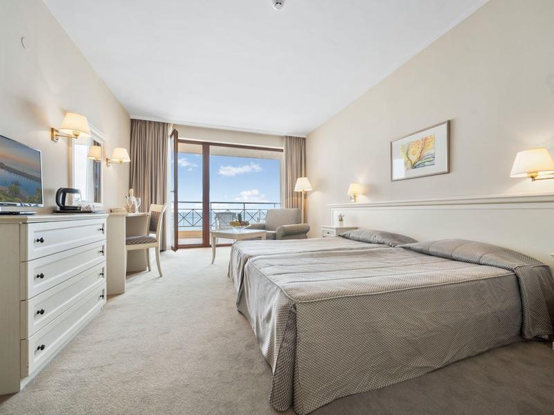 Bright hotel room with two beds, TV, desk, and balcony with sea view.