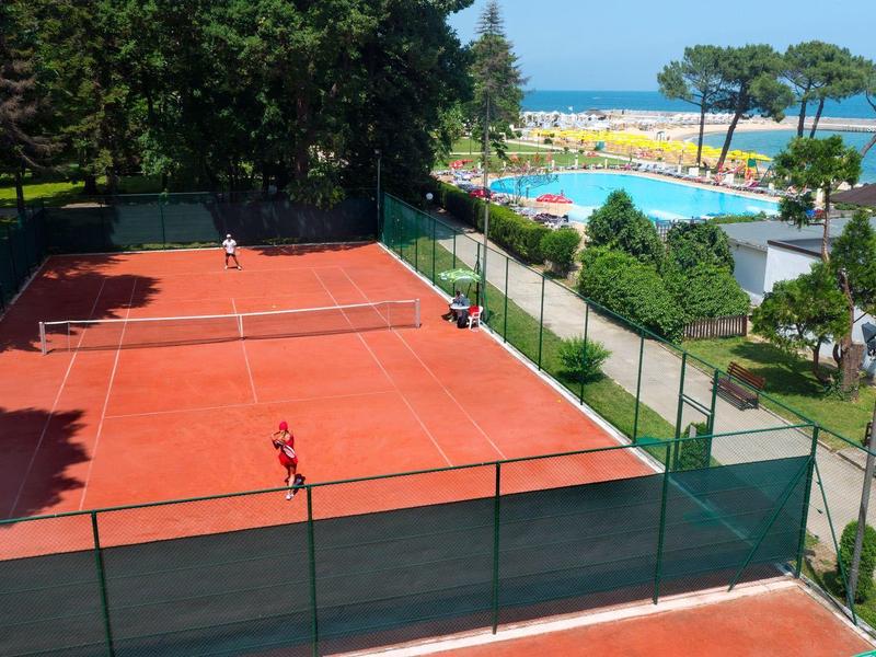 View of red clay tennis courts with sea view and surrounding greenery at a holiday resort.