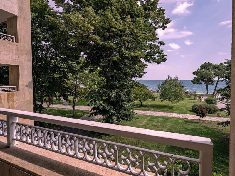 Uitzicht vanaf een balkon met balustrade op een park met bomen en zee aan de horizon.