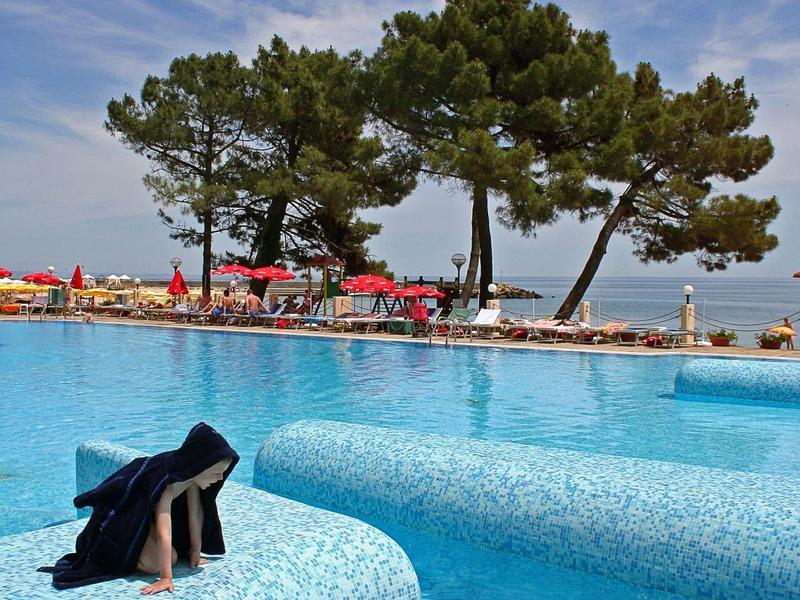 Persoon in blauw zwembad met ligstoelen en bomen aan zee op de achtergrond