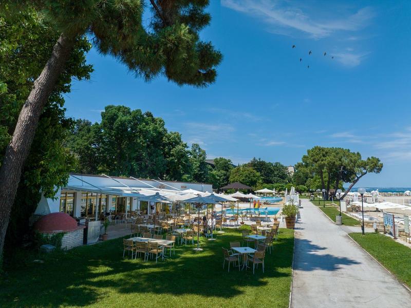 Strandpromenade met tafels, parasols en bomen onder een blauwe lucht.