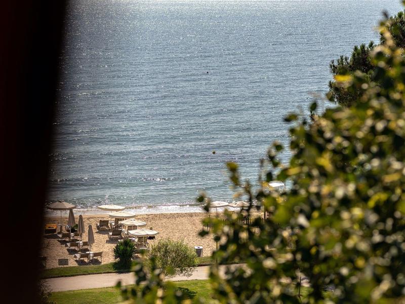 Uitzicht op een strand met parasols en mensen, omgeven door groen en zee op de achtergrond.