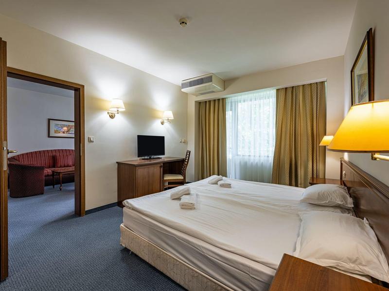 Lichte hotelkamer met tweepersoonsbed, tv, fauteuils en gordijnen bij een raam.