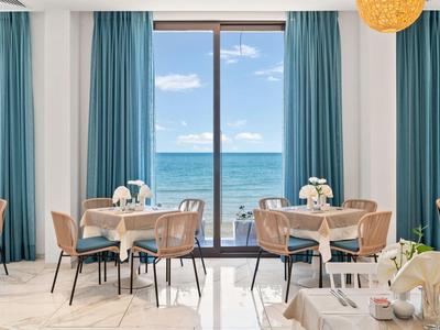 Světlá jídelna s modrými závěsy a výhledem na moře skrz velká okna.