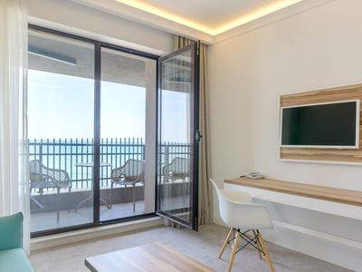 Světly hotelový pokoj s TV, stolem a balkonem s posezením a výhledem na moře.