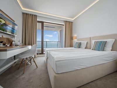 Světle zařízený hotelový pokoj s manželskou postelí a balkonem s výhledem na moře.